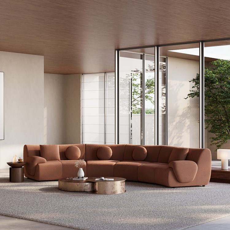 Curved rust-brown sofa with round and square cushions faces two low coffee tables on a pale rug in a bright room with floor-to-ceiling windows.