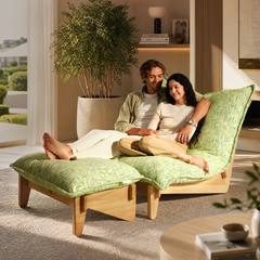 Two smiling people recline together on a green cushioned lounge with a matching footrest in a sunlit room beside a large potted tree.