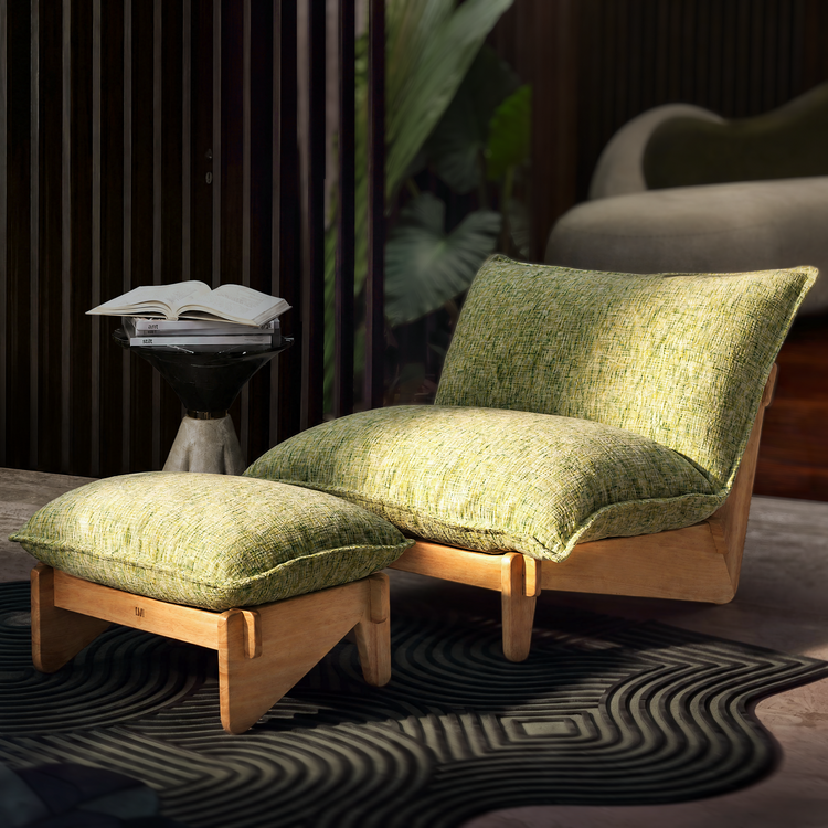 Lounge chair and matching footstool with light brown frames and textured yellow-green cushions on a dark patterned rug, beside a small table holding stacked books and an open book.