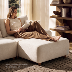 A woman reads a book while reclining barefoot on a cream sofa with her legs stretched across a matching ottoman, beside shelves of books and a sunlit window.