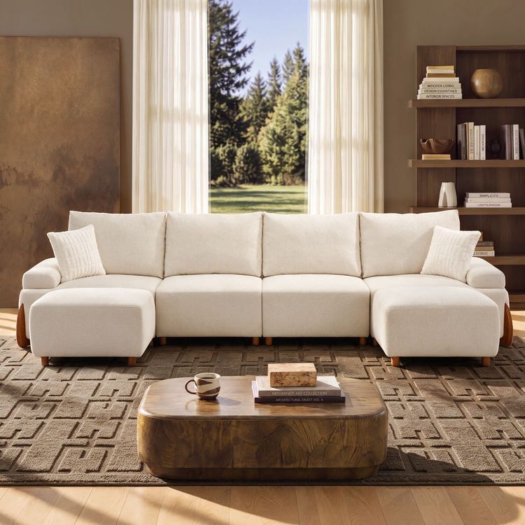 A cream sectional sofa with two ottomans sits in a sunlit living room, with a coffee table, patterned rug, bookshelves, and trees visible through the window.