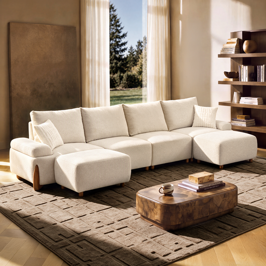 Cream sofa with two matching footstools and throw pillows in a sunlit room with a patterned rug, low brown coffee table, open shelving and tall curtained windows.