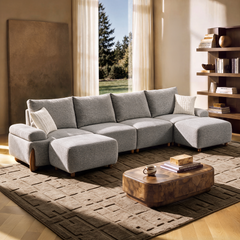 A gray sectional sofa with two ottomans and white cushions sits on a patterned rug beside a wooden coffee table in a sunlit living room.