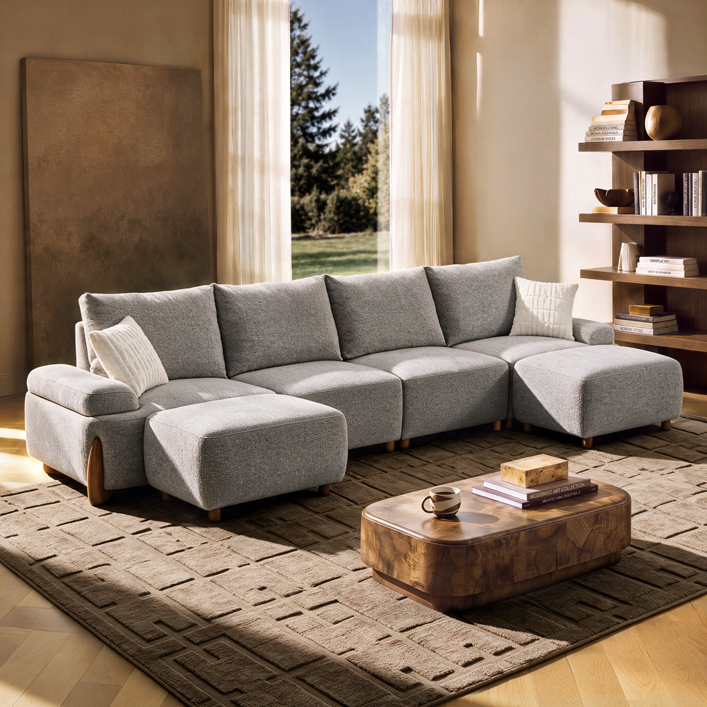A gray sectional sofa with two ottomans and white cushions sits on a patterned rug beside a wooden coffee table in a sunlit living room.