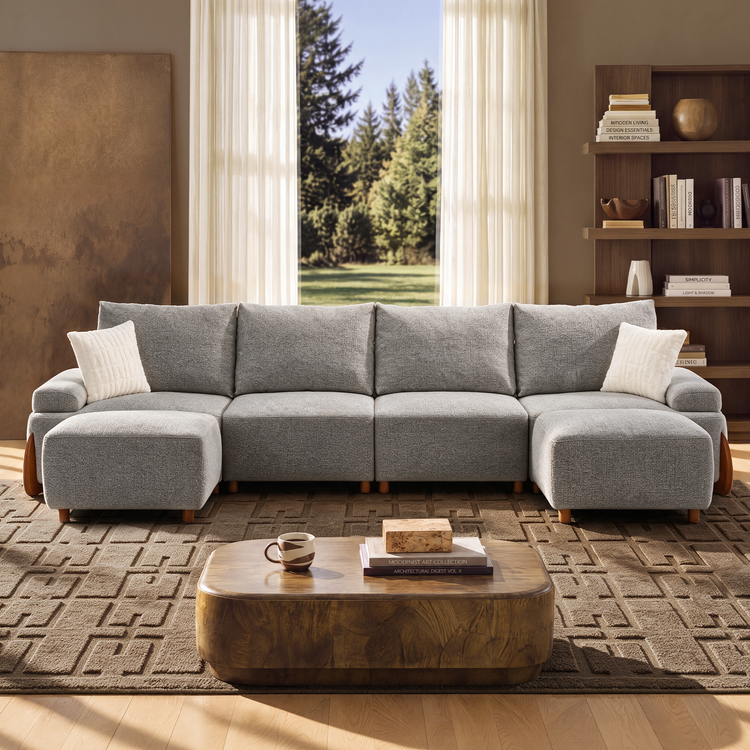 A gray sectional sofa with two white pillows sits in a sunlit living room, facing a wooden coffee table and a window overlooking trees.
