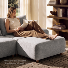 A woman sits barefoot on a gray sectional sofa reading a book beside a wooden shelf, in a sunlit living room.