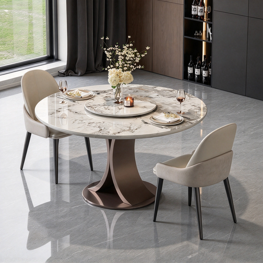 Round dining table with a veined top and sculptural pedestal, set with plates, glasses, flowers and a candle, flanked by two cream chairs near a window and dark cabinets.