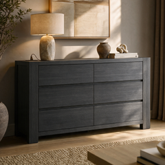 Dark gray six-drawer dresser with visible grain, topped with a beige-shaded lamp, a vase, and books supporting a knot sculpture, beneath framed abstract artwork.