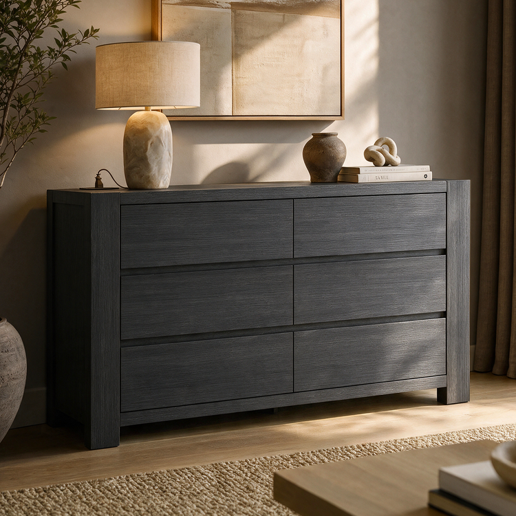 Dark gray six-drawer dresser with visible grain, topped with a beige-shaded lamp, a vase, and books supporting a knot sculpture, beneath framed abstract artwork.