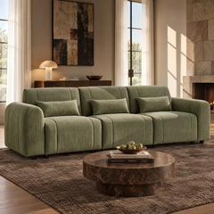 Sage-green sofa with rounded arms, three seats and three matching rectangular cushions, set behind a round brown coffee table in a sunlit living room with a fireplace.
