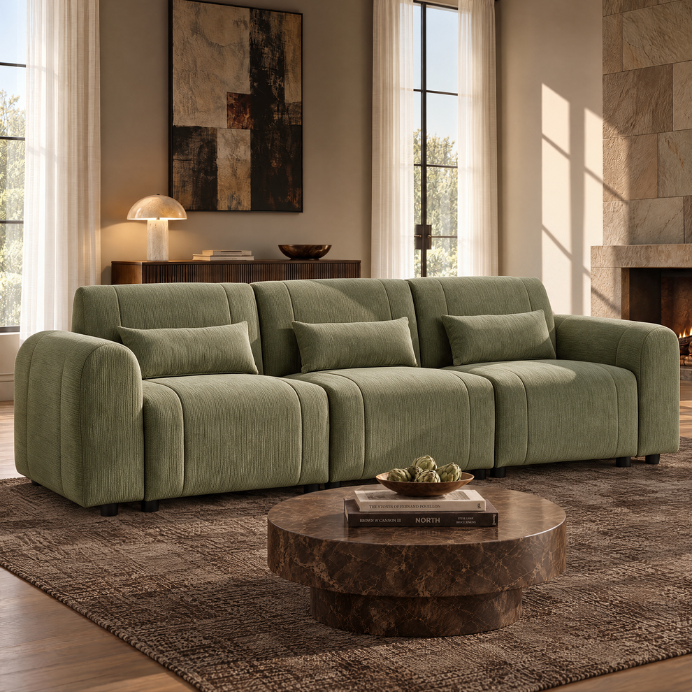 Sage-green sofa with rounded arms, three seats and three matching rectangular cushions, set behind a round brown coffee table in a sunlit living room with a fireplace.
