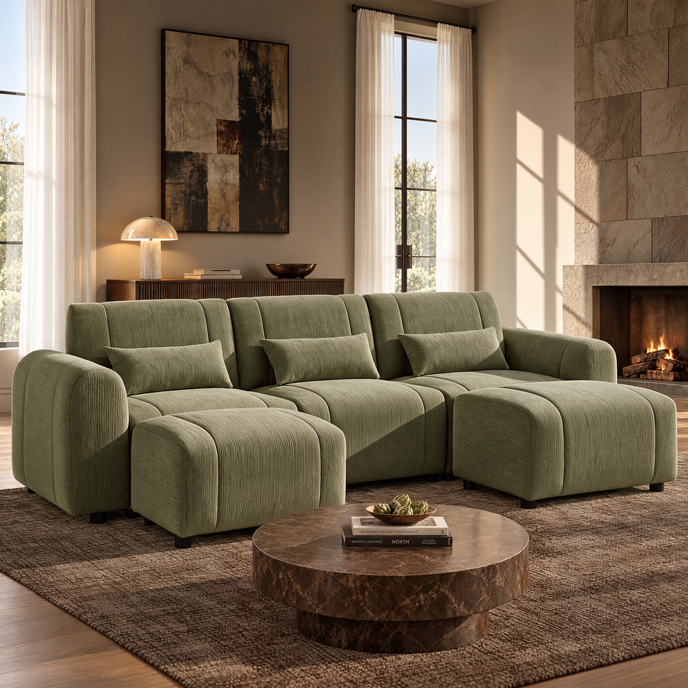 Green upholstered sofa with three matching cushions and two ottomans in a sunlit living room with a round brown coffee table, abstract wall art and a lit fireplace.