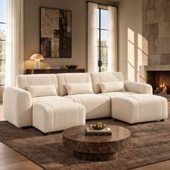 Cream sofa with rounded arms, three back cushions and matching pillows, paired with two ottomans in a sunlit living room with a round brown coffee table and lit fireplace.