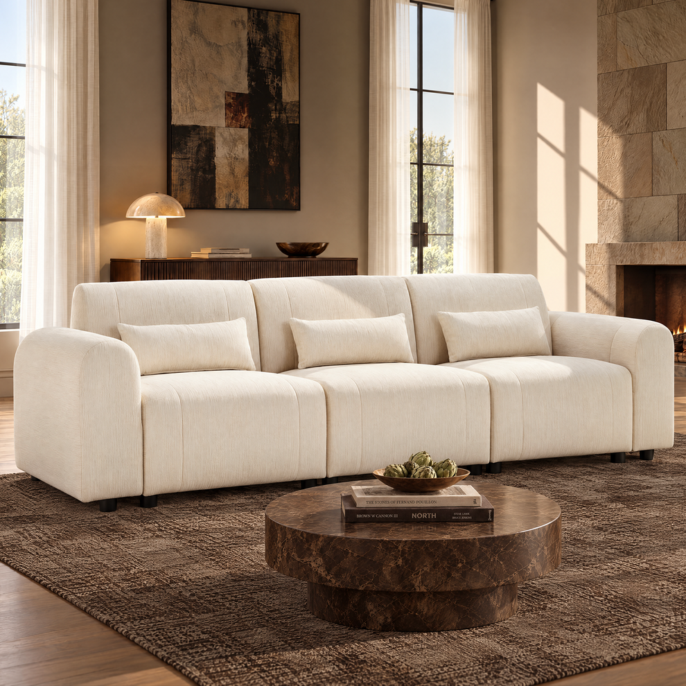 Cream three-seat sofa with rounded arms and three rectangular pillows in a sunlit living room, with a round brown coffee table, textured rug, tall windows and a fireplace.