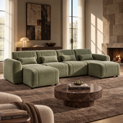 Green upholstered sofa with four matching cushions and two ottomans in a sunlit living room with a round brown coffee table, abstract wall art and a lit fireplace.