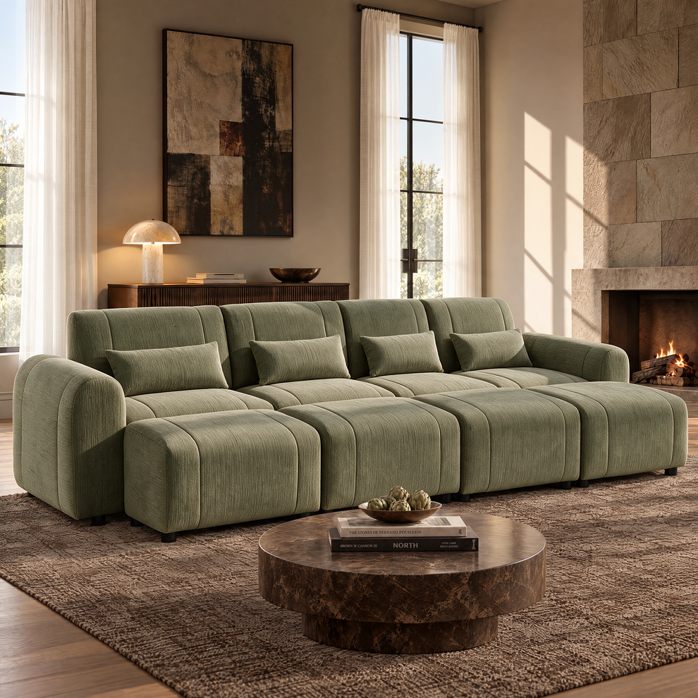Sage-green sofa with rounded arms, four matching cushions and four ottomans in a sunlit living room with a round brown coffee table, abstract wall art and a lit fireplace.