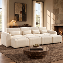 Cream sofa with four matching pillows and four ottomans in a sunlit living room with a round brown coffee table, textured rug, abstract wall art and a lit fireplace.
