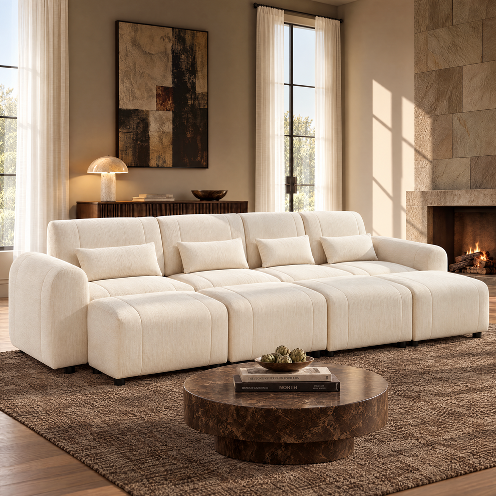 Cream sofa with four matching pillows and four ottomans in a sunlit living room with a round brown coffee table, textured rug, abstract wall art and a lit fireplace.