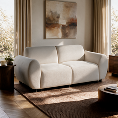 Cream-colored sofa with rounded arms, two seats and short dark legs sits on a brown rug beneath an abstract painting, between curtained windows.