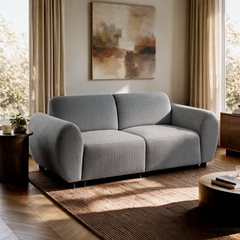 Gray two-seat sofa in a sunlit living room, with abstract wall art, beige curtains, a woven rug, and wooden side tables.