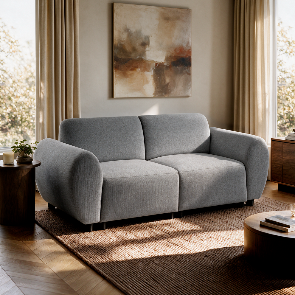 Gray two-seat sofa in a sunlit living room, with abstract wall art, beige curtains, a woven rug, and wooden side tables.