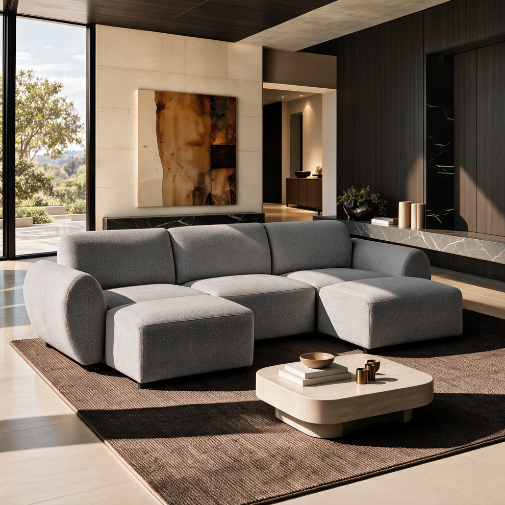 Light gray sofa with rounded arms and two extended front sections on a brown rug, beside a low cream coffee table in a sunlit living room with tall windows and abstract wall art.