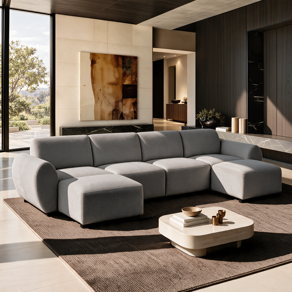 Light gray sectional sofa with rounded arms and two projecting end seats on a brown rug, facing a low coffee table in a sunlit living room with tall windows and abstract wall art.