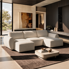 Cream upholstered sofa with rounded arms and two matching ottomans on a brown rug, beside a low coffee table in a sunlit living room with abstract wall art and tall windows.