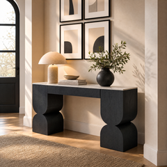 Black sculptural console table styled with a lamp, books, bowl, and leafy vase.