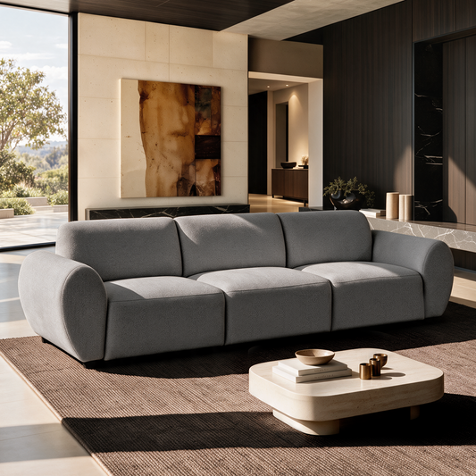 Gray three-seat sofa with rounded arms on a brown rug in a sunlit living room, with a low cream coffee table, abstract wall art and a large window.