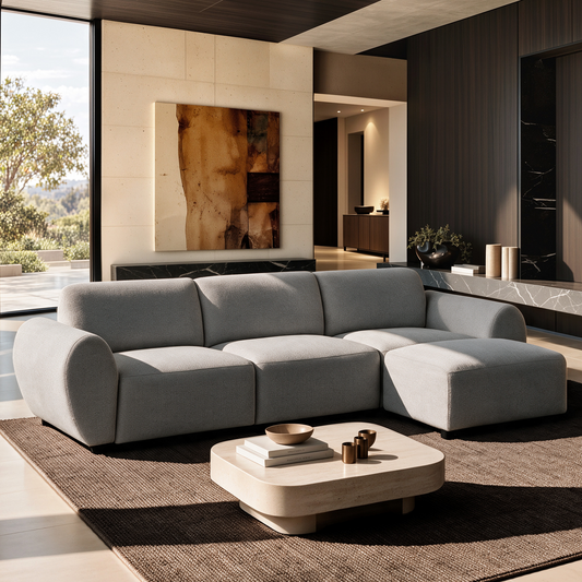 Light gray sectional sofa with rounded arms and a right-side chaise in a sunlit living room with a low cream coffee table, brown rug, abstract wall art and large window.