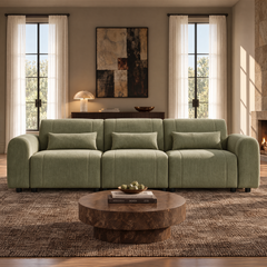 Pale green three-seat sofa with rounded arms and three matching pillows in a living room with a round brown coffee table, abstract wall art, tall curtained windows and a lit fireplace.