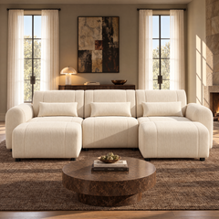 Cream sofa with rounded arms, three back cushions and matching pillows, with two matching ottomans in front, facing a round brown coffee table in a sunlit living room.