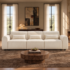 Cream three-seat sofa with rounded arms and three rectangular pillows in a sunlit living room with a round brown coffee table, patterned rug, abstract wall art and sheer curtains.