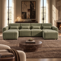 Olive-green sectional sofa with four cushions in a sunlit living room, with a round coffee table, abstract wall art, tall windows, and a stone fireplace.