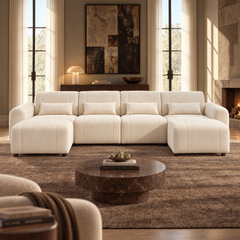 Cream sectional sofa with extended seats at both ends and four matching pillows in a sunlit living room with a round brown coffee table, abstract wall art and a lit fireplace.