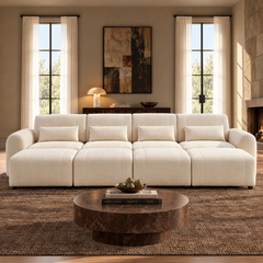 Cream four-seat sofa with rounded arms, four matching pillows and extended footrests in a living room with a round brown coffee table, tall curtained windows and a fireplace.