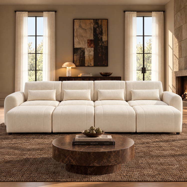 Cream four-seat sofa with rounded arms, four matching pillows and extended footrests in a living room with a round brown coffee table, tall curtained windows and a fireplace.