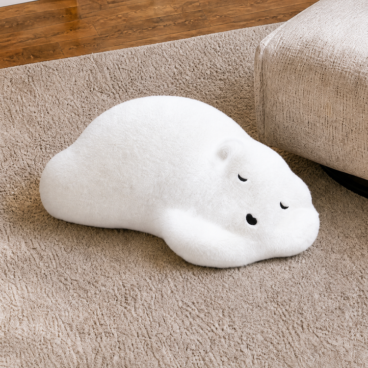 A flattened animal-shaped plush cushion with stitched closed eyes and nose rests on a textured rug beside a sofa