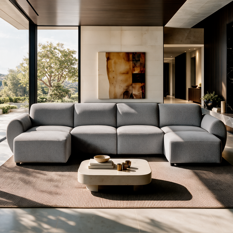 Gray sectional sofa with rounded arms and extended seats at both ends faces a low coffee table in a sunlit room with large windows and abstract wall art.