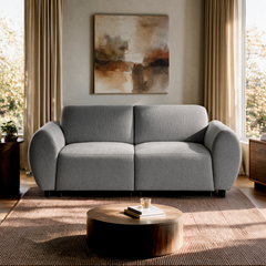 Light gray two-seat sofa with rounded arms beneath an abstract painting, flanked by curtained windows, with a round coffee table on a brown rug in front.