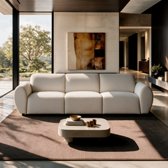 Cream three-seat sofa with rounded arms in a sunlit living room, with a low coffee table, brown rug, abstract wall art and tall windows overlooking trees.