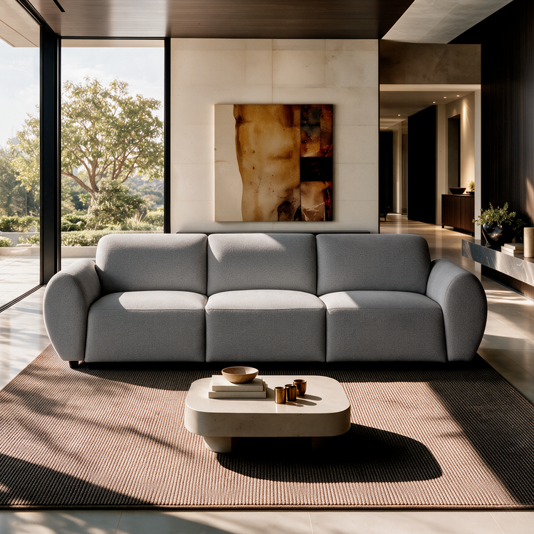 Gray three-seat sofa with rounded arms in a sunlit living room, with a low coffee table on a textured rug, abstract wall art and tall windows overlooking trees.