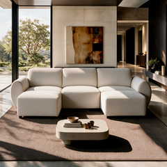 Cream sofa with rounded arms and two extended end seats in a sunlit living room with a low coffee table, textured rug, abstract wall art and floor-to-ceiling windows.