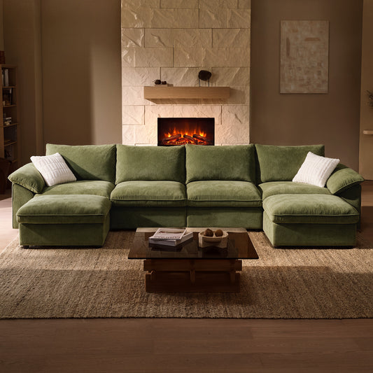 Green sofa with chaise extensions at both ends and two white pillows, facing a low coffee table on a beige rug, with a lit fireplace behind it.