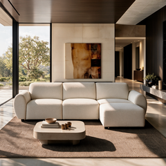 Cream sectional sofa with rounded arms and a right-side chaise in a sunlit living room, with a low coffee table, textured rug, abstract wall art and floor-to-ceiling windows.