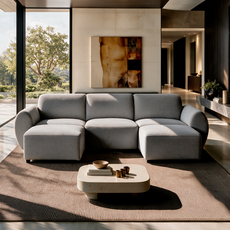 Gray sofa with rounded arms and two projecting end seats in a sunlit living room, with a low coffee table, textured rug, abstract wall art and tall windows overlooking trees.