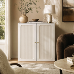 White storage cabinet with fluted double doors and gold-tone knobs beside a living room window