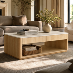 A light wood coffee table with a pale top and open shelf stands between sofas; books and a woven basket sit on the shelf, with a vase of leafy branches on top.