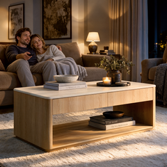 A wooden coffee table with a light-colored top, drawer, and lower shelf stands in a warmly lit living room with books, bowls, a candle, and a vase of foliage.